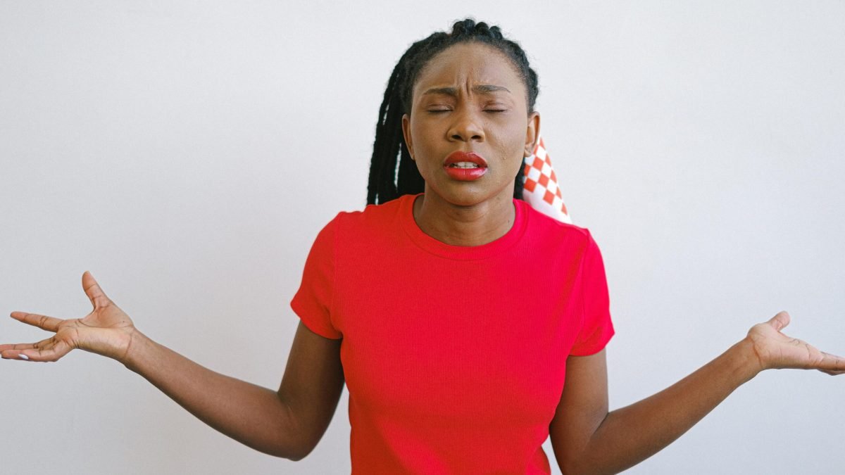 Portrait of a woman in a red shirt expressing confusion with her eyes closed and arms out. Ideal for emotion concepts.