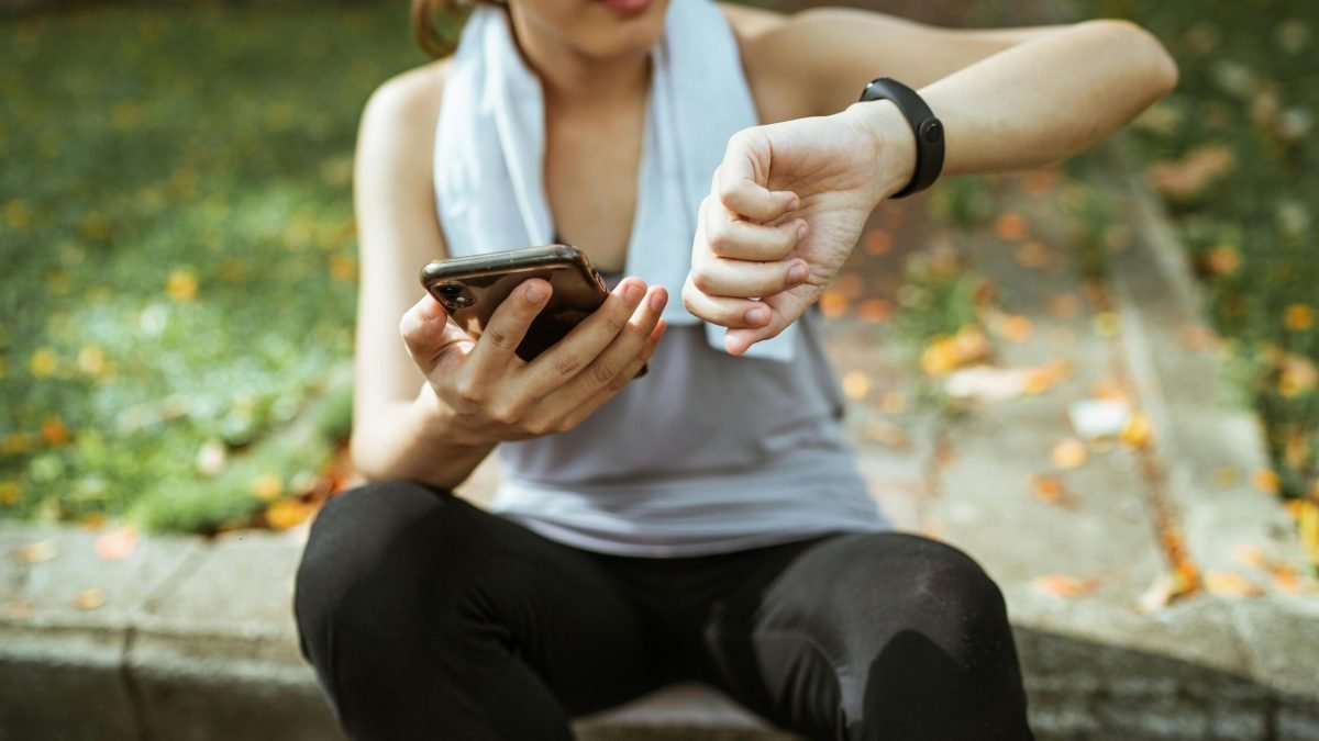 Anonymous female in sportswear using smartphone and smart watch after exercising on sports ground