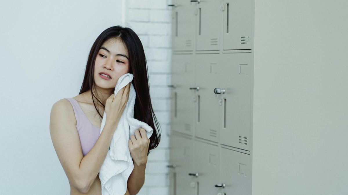 Woman in a locker room drying off with a white towel, next to lockers.