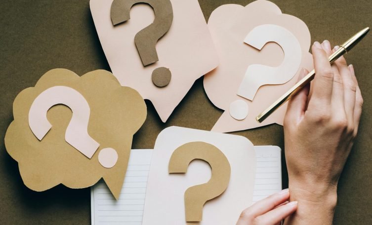 Hands holding pen next to paper question marks on notebook, symbolizing inquiry and creativity.