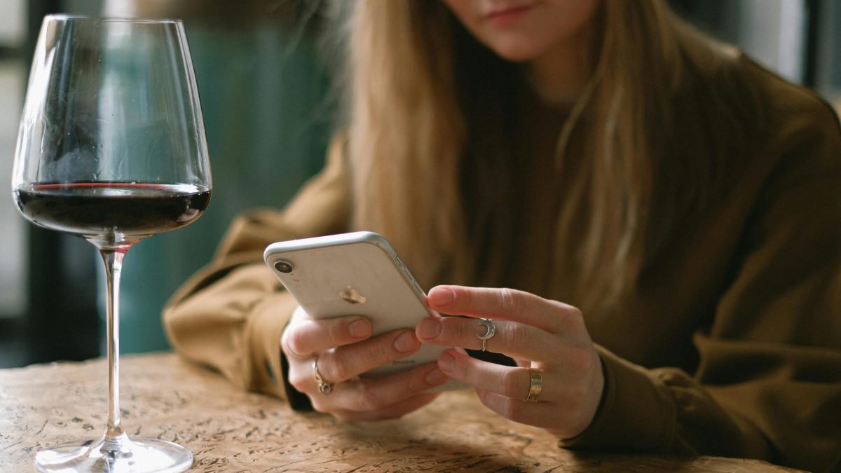 A woman is relaxing indoors, using her smartphone with a glass of red wine on the table.