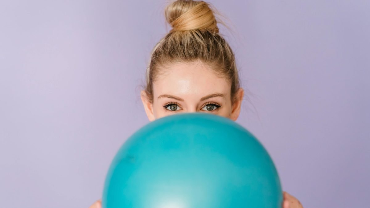 Cheerful anonymous female demonstrating blue shiny gym ball in hands on purple background of studio