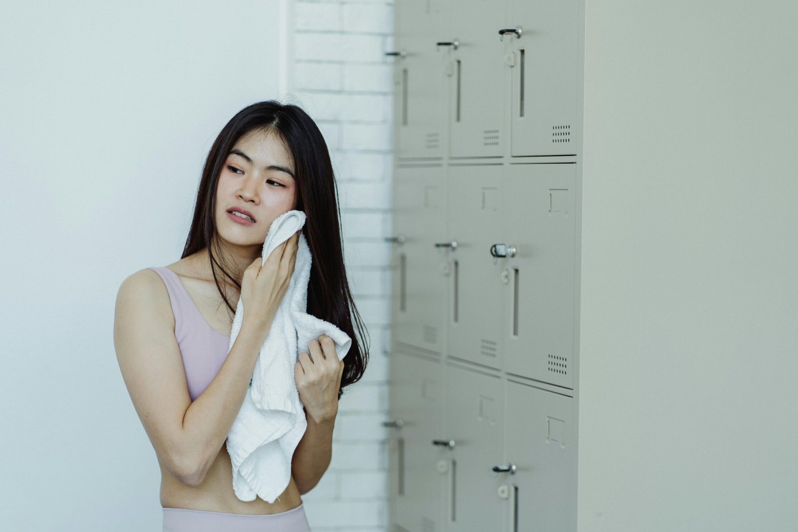 Woman in a locker room drying off with a white towel, next to lockers.