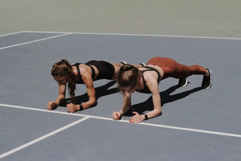 Two women in activewear doing plank exercises on an outdoor sports court.